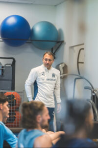 Coach in white training top speaks to athletes in a gym, with blue exercise balls on a shelf behind him.